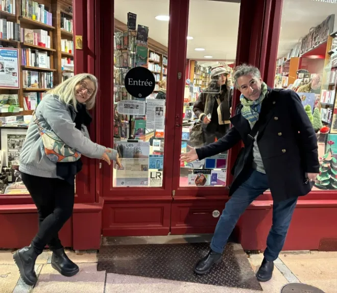 Deux personnes souriantes devant l’entrée d’une librairie, montrant la porte entourée de vitrines remplies de livres.