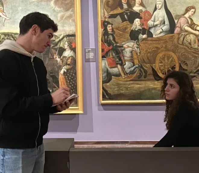 Deux personnes dans un musée : l’une prend des notes sur un carnet tandis que l’autre est assise devant de grands tableaux anciens.
