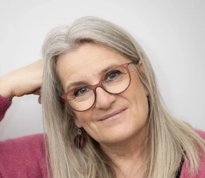 Portrait d’une personne aux cheveux gris et lunettes rouges, souriant légèrement devant un fond clair.